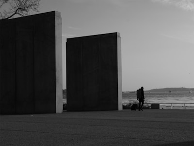 The image depicts a solitary figure walking with a suitcase near two large, monolithic structures by a waterfront. The background shows a calm body of water with distant land and a few visible boats, under a partly cloudy sky. The setting appears peaceful and somewhat isolated.