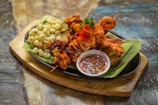 A platter featuring grilled seafood skewers including shrimp and octopus, served on a banana leaf with a side of small white corns and a dish of dipping sauce. The arrangement is colorful and garnished with parsley and red pepper.