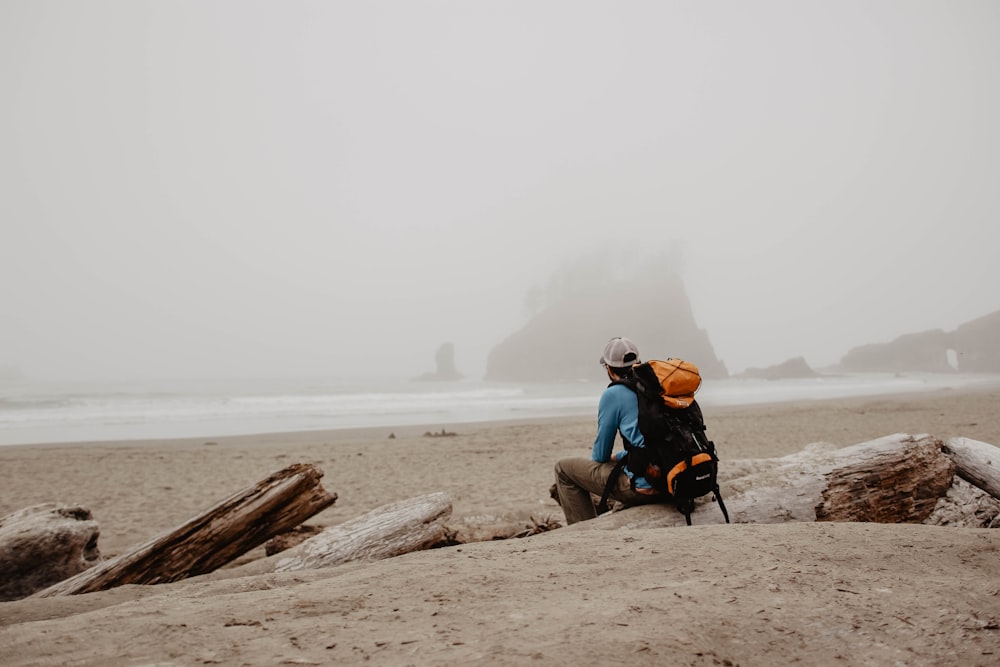 homem vestindo mochila de caminhada laranja e preta sentado em log na costa olhando para a distância durante o dia