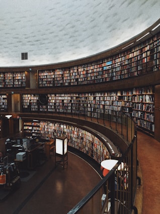 A large, circular library space with multiple levels of bookshelves filled with numerous books. The architectural design features a high, dome-like ceiling and a warm ambiance. There are desks and computers in the foreground, suggesting a space for reading and research.