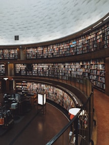 A large, circular library space with multiple levels of bookshelves filled with numerous books. The architectural design features a high, dome-like ceiling and a warm ambiance. There are desks and computers in the foreground, suggesting a space for reading and research.
