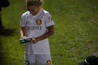 A person wearing a white sports jersey, holding a book or magazine and a pen, appears to be signing an autograph. The sports jersey has logos and text, indicating it may be part of a team uniform. The scene takes place on a grassy field with another person partially visible kneeling on the ground.