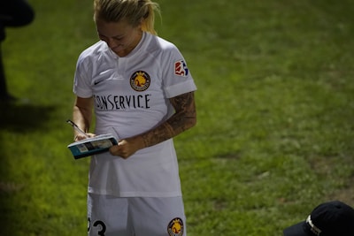 A person wearing a white sports jersey, holding a book or magazine and a pen, appears to be signing an autograph. The sports jersey has logos and text, indicating it may be part of a team uniform. The scene takes place on a grassy field with another person partially visible kneeling on the ground.