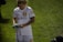 A person wearing a white sports jersey, holding a book or magazine and a pen, appears to be signing an autograph. The sports jersey has logos and text, indicating it may be part of a team uniform. The scene takes place on a grassy field with another person partially visible kneeling on the ground.