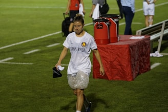 A person wearing a white sports jersey with 'CONSERVICE' written on it and ice packs wrapped around both knees is walking on a grass field. The background includes a red table with large cooler jugs on it and several other people, some blurred, standing around. The person appears to be holding a black cap and a cloth.