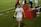 A person wearing a white sports jersey with 'CONSERVICE' written on it and ice packs wrapped around both knees is walking on a grass field. The background includes a red table with large cooler jugs on it and several other people, some blurred, standing around. The person appears to be holding a black cap and a cloth.