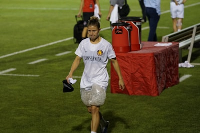A person wearing a white sports jersey with 'CONSERVICE' written on it and ice packs wrapped around both knees is walking on a grass field. The background includes a red table with large cooler jugs on it and several other people, some blurred, standing around. The person appears to be holding a black cap and a cloth.