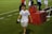 A person wearing a white sports jersey with 'CONSERVICE' written on it and ice packs wrapped around both knees is walking on a grass field. The background includes a red table with large cooler jugs on it and several other people, some blurred, standing around. The person appears to be holding a black cap and a cloth.