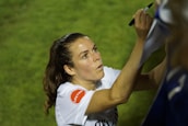 A person with long hair is signing something with a black marker. They are wearing a white shirt with a red patch on the sleeve. The background is grassy and out of focus.