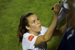 A person with long hair is signing something with a black marker. They are wearing a white shirt with a red patch on the sleeve. The background is grassy and out of focus.