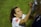 A person with long hair is signing something with a black marker. They are wearing a white shirt with a red patch on the sleeve. The background is grassy and out of focus.