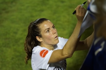 A person with long hair is signing something with a black marker. They are wearing a white shirt with a red patch on the sleeve. The background is grassy and out of focus.