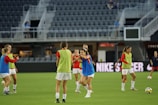 A group of female soccer players are on a field, wearing training bibs over their uniforms. They are practicing with a soccer ball, engaged in a passing drill. The stadium appears empty except for a few spectators and staff. The players are focused and in motion, showing dynamic movement.