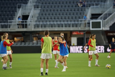 A group of female soccer players are on a field, wearing training bibs over their uniforms. They are practicing with a soccer ball, engaged in a passing drill. The stadium appears empty except for a few spectators and staff. The players are focused and in motion, showing dynamic movement.