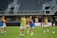 A group of female soccer players are on a field, wearing training bibs over their uniforms. They are practicing with a soccer ball, engaged in a passing drill. The stadium appears empty except for a few spectators and staff. The players are focused and in motion, showing dynamic movement.