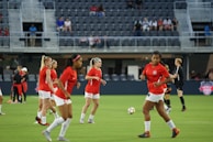 A group of female soccer players in red jerseys and white shorts are on a soccer field, engaging in warm-up exercises or practice. The players are in various positions, some jogging and others preparing to kick a ball. A soccer ball is visible on the ground, and a referee in black uniform is on the field as well. The background shows empty stadium seats with some spectators visible.