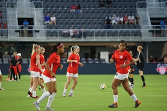A group of female soccer players in red jerseys and white shorts are on a soccer field, engaging in warm-up exercises or practice. The players are in various positions, some jogging and others preparing to kick a ball. A soccer ball is visible on the ground, and a referee in black uniform is on the field as well. The background shows empty stadium seats with some spectators visible.