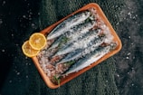 Fresh sardines are neatly arranged on a wooden tray covered with coarse sea salt. Lemon slices and sprigs of fresh herbs complement the fish, and a fishing net is partially visible beneath the tray.