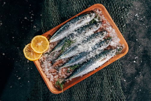 Fresh sardines are neatly arranged on a wooden tray covered with coarse sea salt. Lemon slices and sprigs of fresh herbs complement the fish, and a fishing net is partially visible beneath the tray.