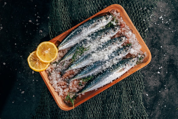 Fresh sardines are neatly arranged on a wooden tray covered with coarse sea salt. Lemon slices and sprigs of fresh herbs complement the fish, and a fishing net is partially visible beneath the tray.