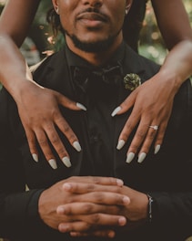 A person dressed in a formal black suit with a bow tie is seated, showing only the lower face and hands folded in front. Another person's hands are gently placed on their shoulders, adorned with an engagement ring and light-colored nail polish.