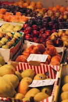 A variety of fresh fruits are neatly arranged in boxes at a market. The selection includes citrus fruits, apples, plums, figs, and peaches. Each box is labeled with handwritten signs displaying prices or names. The scene is colorful and vibrant, highlighting the abundance of fresh produce available.