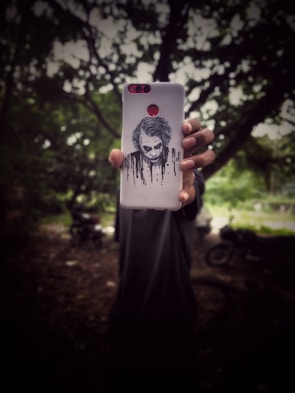 A person is holding a smartphone case with an artistic design of a sinister-looking face with dark streaks. The background is a blurred outdoor setting with trees and a motorcycle partially visible, creating a mysterious atmosphere.