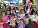 A group of children and two adults wearing traditional clothing, holding colorful books and toys, in an outdoor setting. The scene is lively with a market or fair environment in the background, featuring tents and a parked pickup truck. The adults and children are smiling, and there are trees providing some shade.