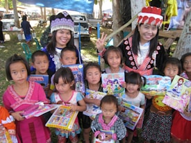 A group of children and two adults wearing traditional clothing, holding colorful books and toys, in an outdoor setting. The scene is lively with a market or fair environment in the background, featuring tents and a parked pickup truck. The adults and children are smiling, and there are trees providing some shade.