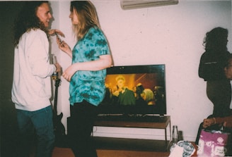 Two people are standing close together in a room, interacting and smiling with each other. One person is holding a drink, while the other appears to be mid-conversation. Behind them, a television displays a scene with multiple people. Additional individuals are seated on the right side of the image. Various objects, including a guitar, bottles, and snacks, are visible in the room.
