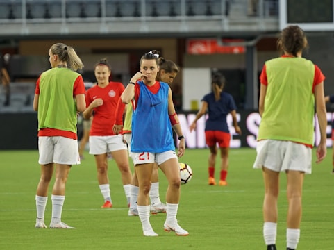 Several female soccer players are on a field during a practice session. They are wearing sports gear, including shirts, shorts, and training vests. Some players are conversing while one is holding a soccer ball. The players appear focused and ready, as they prepare for their practice or match.