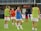 Several female soccer players are on a field during a practice session. They are wearing sports gear, including shirts, shorts, and training vests. Some players are conversing while one is holding a soccer ball. The players appear focused and ready, as they prepare for their practice or match.