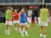 Several female soccer players are on a field during a practice session. They are wearing sports gear, including shirts, shorts, and training vests. Some players are conversing while one is holding a soccer ball. The players appear focused and ready, as they prepare for their practice or match.