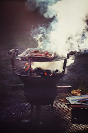 A barbecue grill is set up outdoors, with a metal grate holding meat that is being cooked over hot coals. Thick white smoke billows up from the grill, partially obscuring the meat. The setup is placed on a perforated metal surface, and a disposable aluminum tray is visible beside the grill.