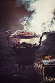 A barbecue grill is set up outdoors, with a metal grate holding meat that is being cooked over hot coals. Thick white smoke billows up from the grill, partially obscuring the meat. The setup is placed on a perforated metal surface, and a disposable aluminum tray is visible beside the grill.