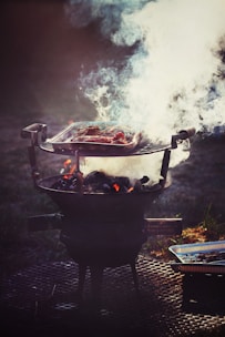 A barbecue grill is set up outdoors, with a metal grate holding meat that is being cooked over hot coals. Thick white smoke billows up from the grill, partially obscuring the meat. The setup is placed on a perforated metal surface, and a disposable aluminum tray is visible beside the grill.