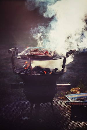 A barbecue grill is set up outdoors, with a metal grate holding meat that is being cooked over hot coals. Thick white smoke billows up from the grill, partially obscuring the meat. The setup is placed on a perforated metal surface, and a disposable aluminum tray is visible beside the grill.