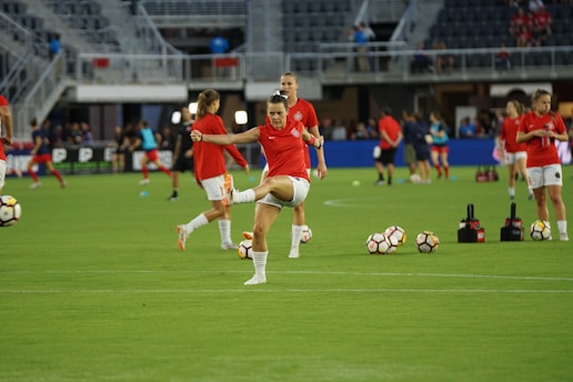 A group of soccer players in red jerseys and white shorts are practicing on a green field. One player in the foreground is stretching their leg, while others are engaged in warming up or training exercises. There are soccer balls scattered on the ground, and the setting appears to be a large sports stadium with empty stands in the background.