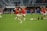 A group of soccer players in red jerseys and white shorts are practicing on a green field. One player in the foreground is stretching their leg, while others are engaged in warming up or training exercises. There are soccer balls scattered on the ground, and the setting appears to be a large sports stadium with empty stands in the background.
