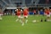 A group of soccer players in red jerseys and white shorts are practicing on a green field. One player in the foreground is stretching their leg, while others are engaged in warming up or training exercises. There are soccer balls scattered on the ground, and the setting appears to be a large sports stadium with empty stands in the background.