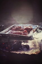 A metal tray filled with seasoned meat pieces is positioned on a charcoal grill emitting gentle smoke. The atmosphere is dimly lit, creating a rustic and warm ambiance.