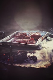 A metal tray filled with seasoned meat pieces is positioned on a charcoal grill emitting gentle smoke. The atmosphere is dimly lit, creating a rustic and warm ambiance.