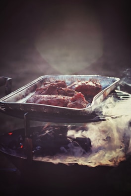 A metal tray filled with seasoned meat pieces is positioned on a charcoal grill emitting gentle smoke. The atmosphere is dimly lit, creating a rustic and warm ambiance.