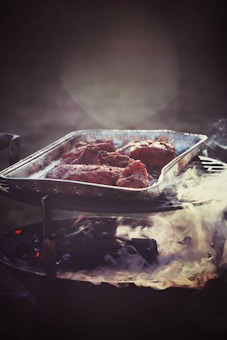 A metal tray filled with seasoned meat pieces is positioned on a charcoal grill emitting gentle smoke. The atmosphere is dimly lit, creating a rustic and warm ambiance.