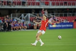 A soccer player in a red jersey prepares to kick a soccer ball on a green field. The background features spectators sitting in red seats, and a sign displaying 'Sports Medicine' is visible.