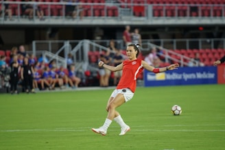 A soccer player in a red jersey prepares to kick a soccer ball on a green field. The background features spectators sitting in red seats, and a sign displaying 'Sports Medicine' is visible.