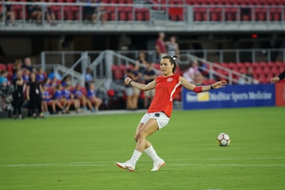 A soccer player in a red jersey prepares to kick a soccer ball on a green field. The background features spectators sitting in red seats, and a sign displaying 'Sports Medicine' is visible.
