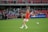 A soccer player in a red jersey prepares to kick a soccer ball on a green field. The background features spectators sitting in red seats, and a sign displaying 'Sports Medicine' is visible.
