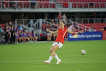 A soccer player in a red jersey prepares to kick a soccer ball on a green field. The background features spectators sitting in red seats, and a sign displaying 'Sports Medicine' is visible.