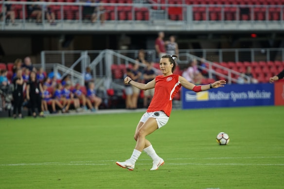 A soccer player in a red jersey prepares to kick a soccer ball on a green field. The background features spectators sitting in red seats, and a sign displaying 'Sports Medicine' is visible.